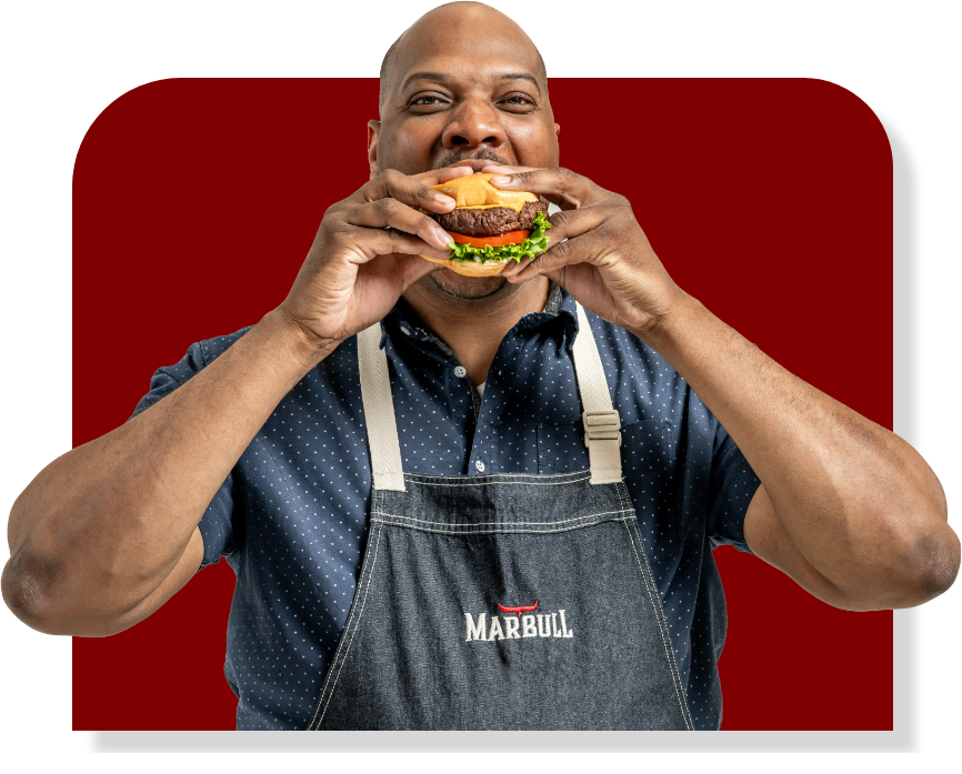 An African America male wearing a blue apron is eating a Marbull wagyu burger.